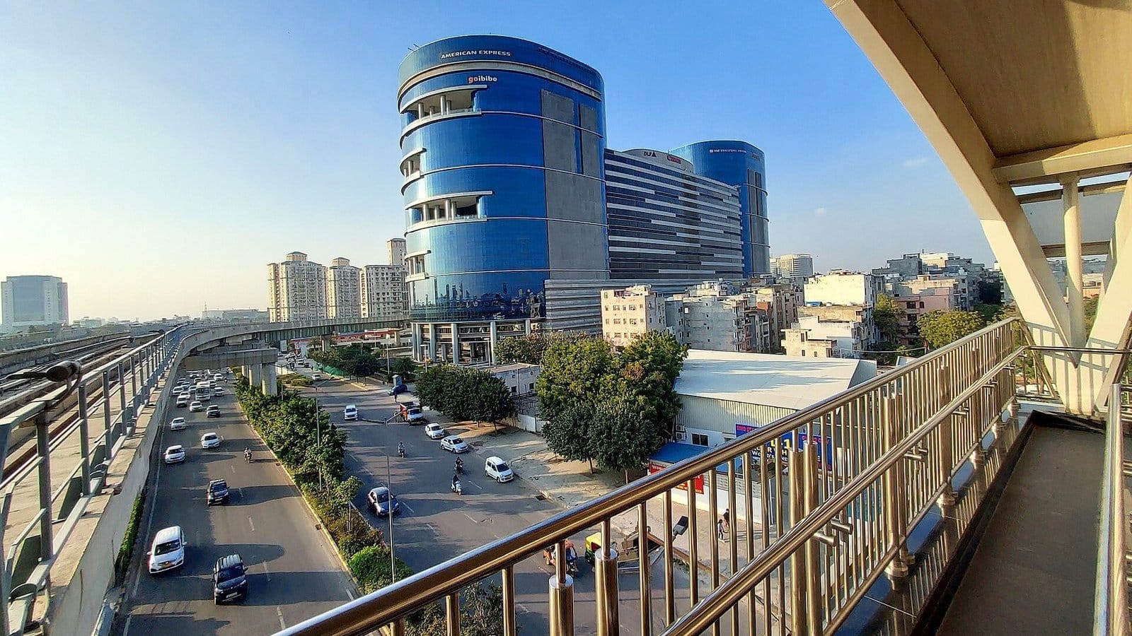 The rounded blue-glass DLF Epitome office building in DLF Phase 3, seen from a raised metro walkway with cars on the road below and apartment towers behind