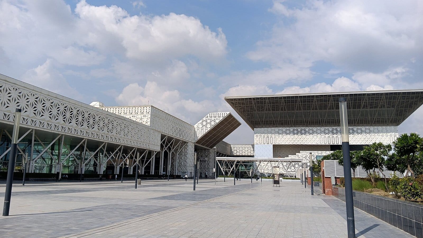 Yashobhoomi convention centre in Dwarka Sector 25, with white lattice facades, a wide cantilevered roof and a paved forecourt under a cloudy blue sky