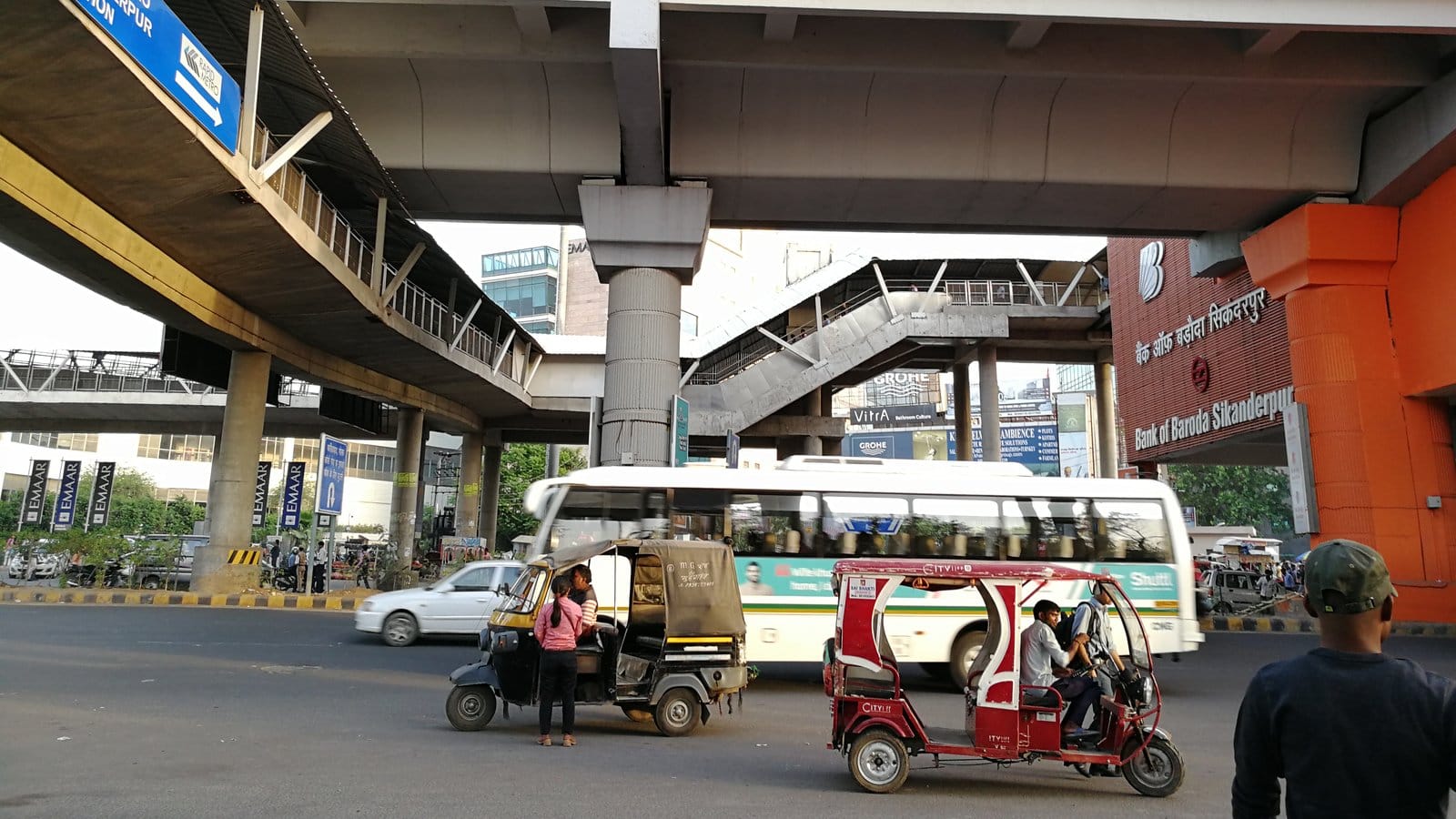 An auto-rickshaw, an e-rickshaw, a car and a white bus on the road under the Sikanderpur metro interchange in Gurgaon, at the northern end of Golf Course Road, in April 2018
