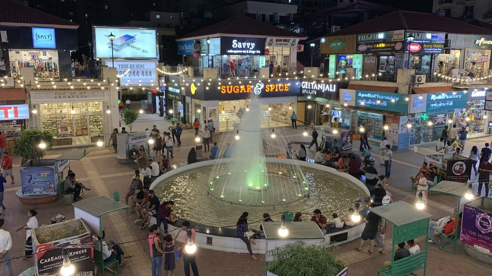 The lit fountain in the middle of DLF Galleria market at night, ringed by people, with Sugar & Spice, Geetanjali Salon and Apollo Pharmacy shopfronts behind it