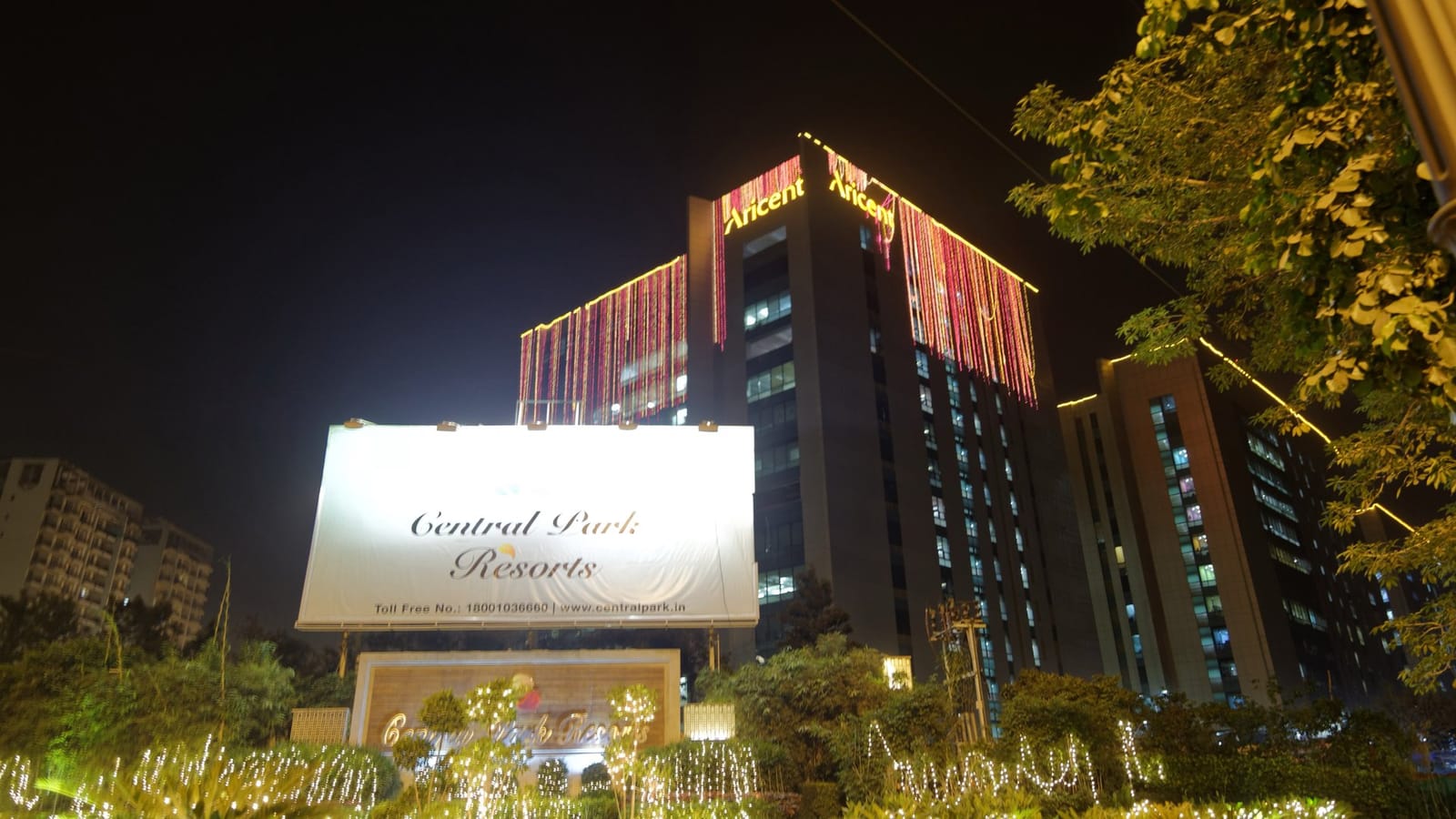 The Central Park Resorts hoarding and entrance garden in Sector 48, Gurgaon, strung with Diwali lights at night, with a lit office tower behind