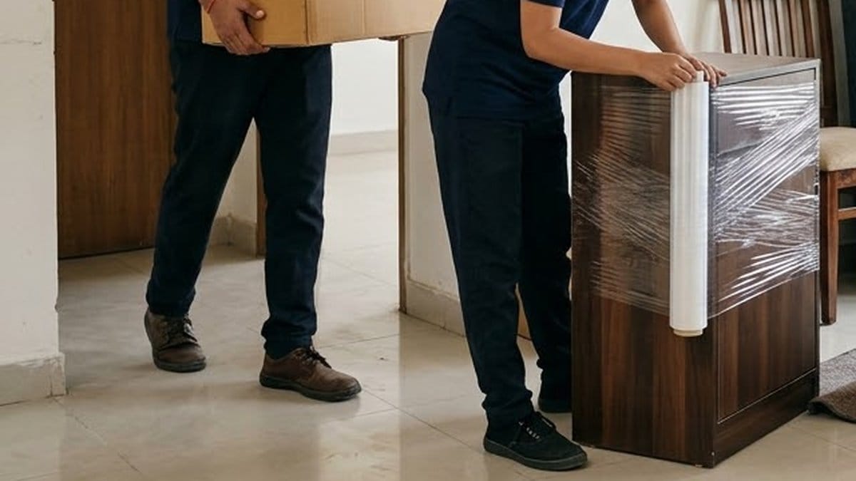 A packer in a navy uniform stretch-wrapping a wooden cabinet indoors while a colleague carries a carton past on a tiled floor
