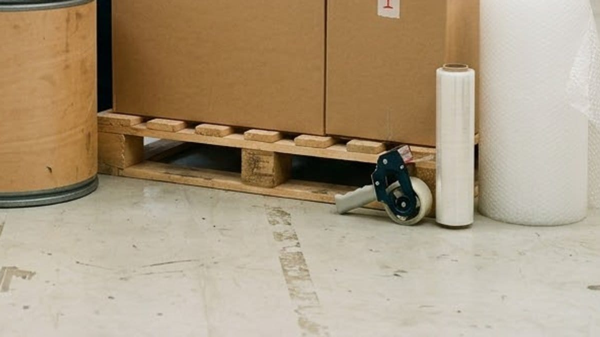 Cartons on a wooden pallet beside a fibre drum, a tape dispenser, a roll of stretch film and a tall roll of bubble wrap on a concrete floor