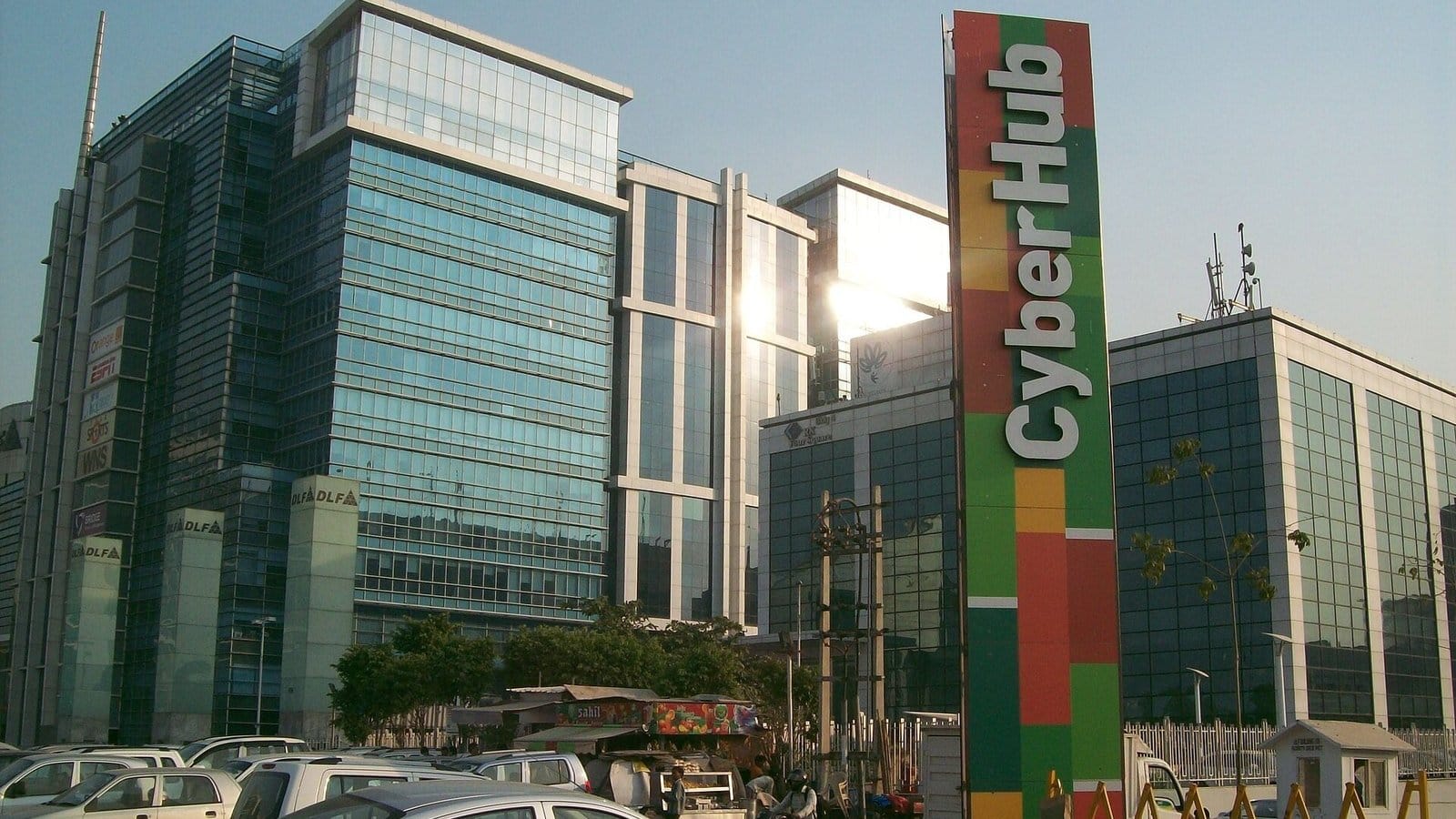 Glass office towers at DLF Cyber City in Gurgaon behind a tall red, green and yellow CyberHub sign, with cars parked in the foreground