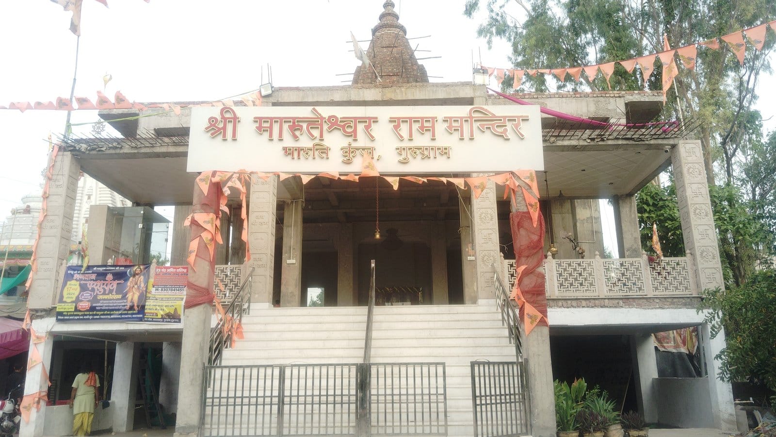 Front of Shree Maruteshwar Ram Mandir in Maruti Kunj, Gurugram, with its Hindi name board above a white staircase, a metal gate and strings of saffron flags