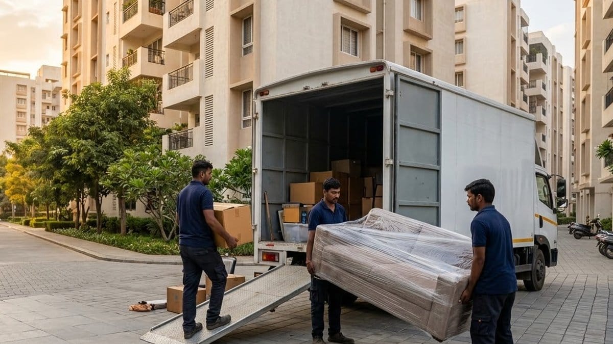 Three movers in navy shirts unload a shrink-wrapped sofa and cartons from a white truck down a ramp on a paved road between apartment towers