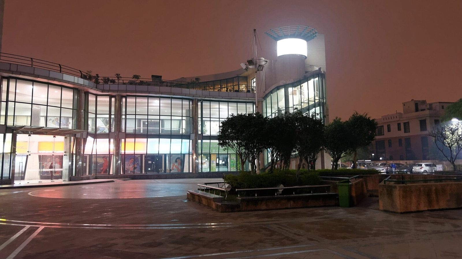 Vipul Arcade in Sushant Lok at dusk: a curved, glass-fronted commercial building with a lit round tower, trees and a paved plaza in front