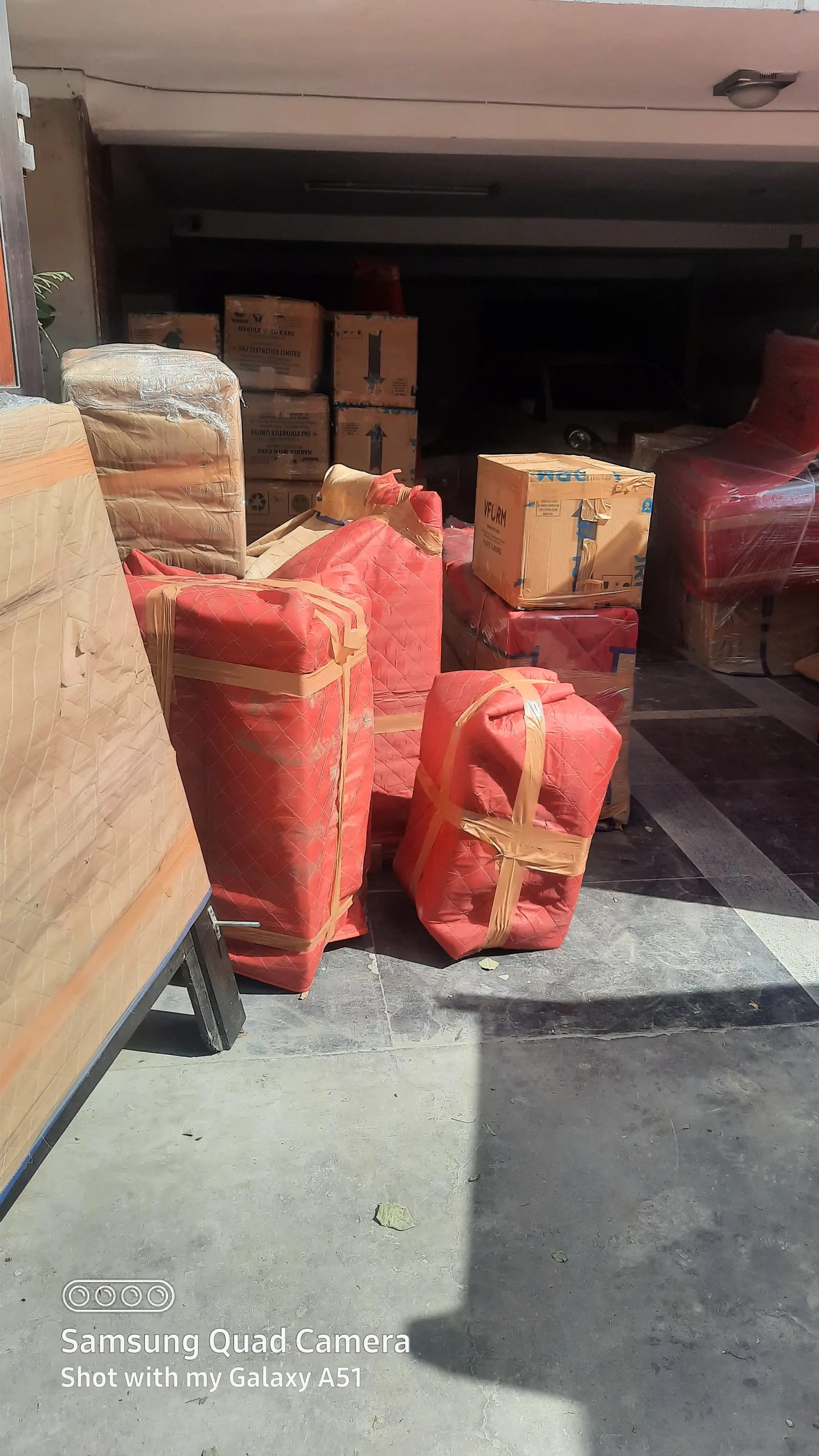 Furniture wrapped in red quilted covers and taped, with sealed cartons behind, in a building’s ground-floor parking