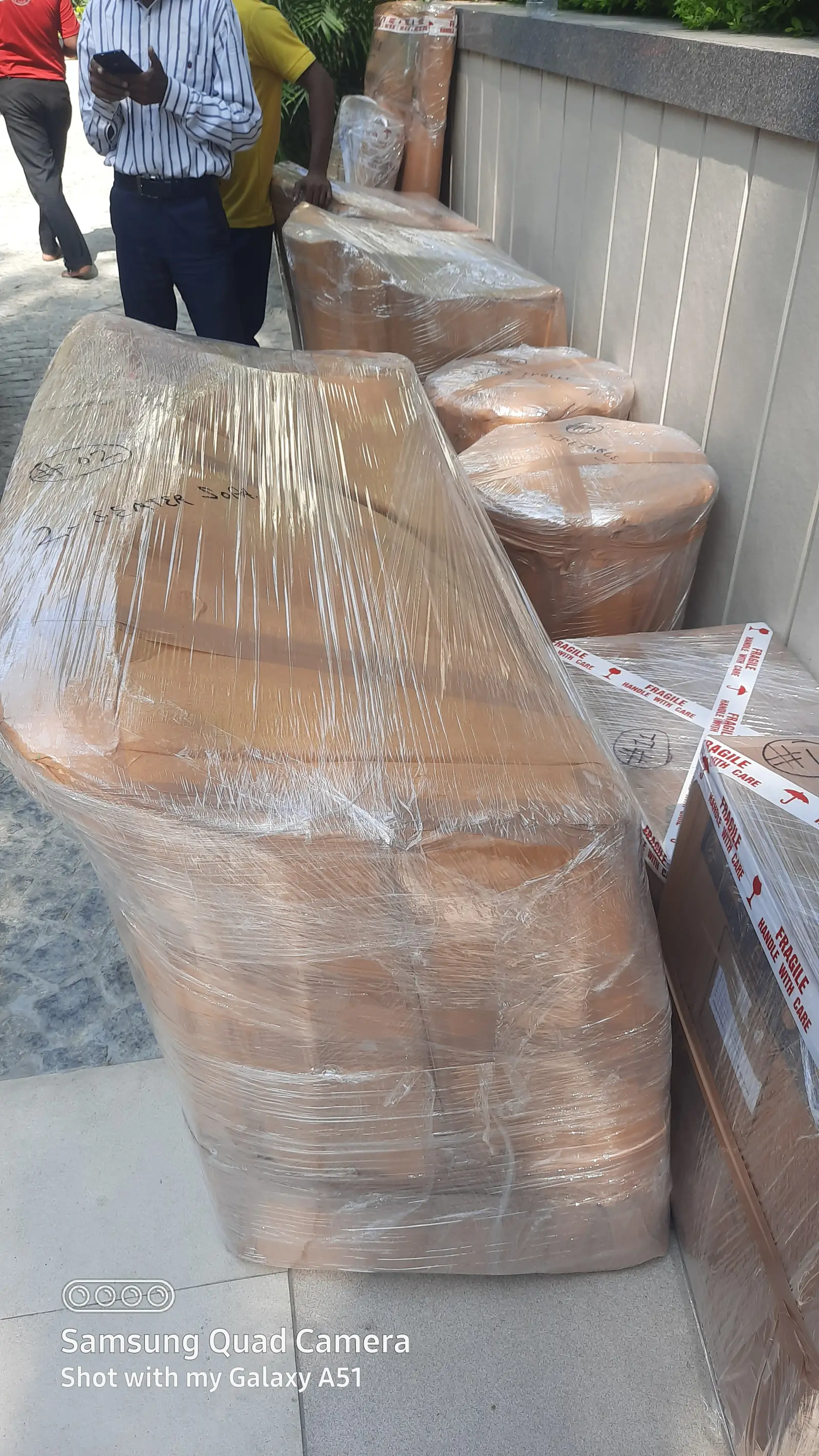 Two-seater sofa and round side tables wrapped in carton sheets and stretch film, lined up on a pavement next to FRAGILE-taped cartons
