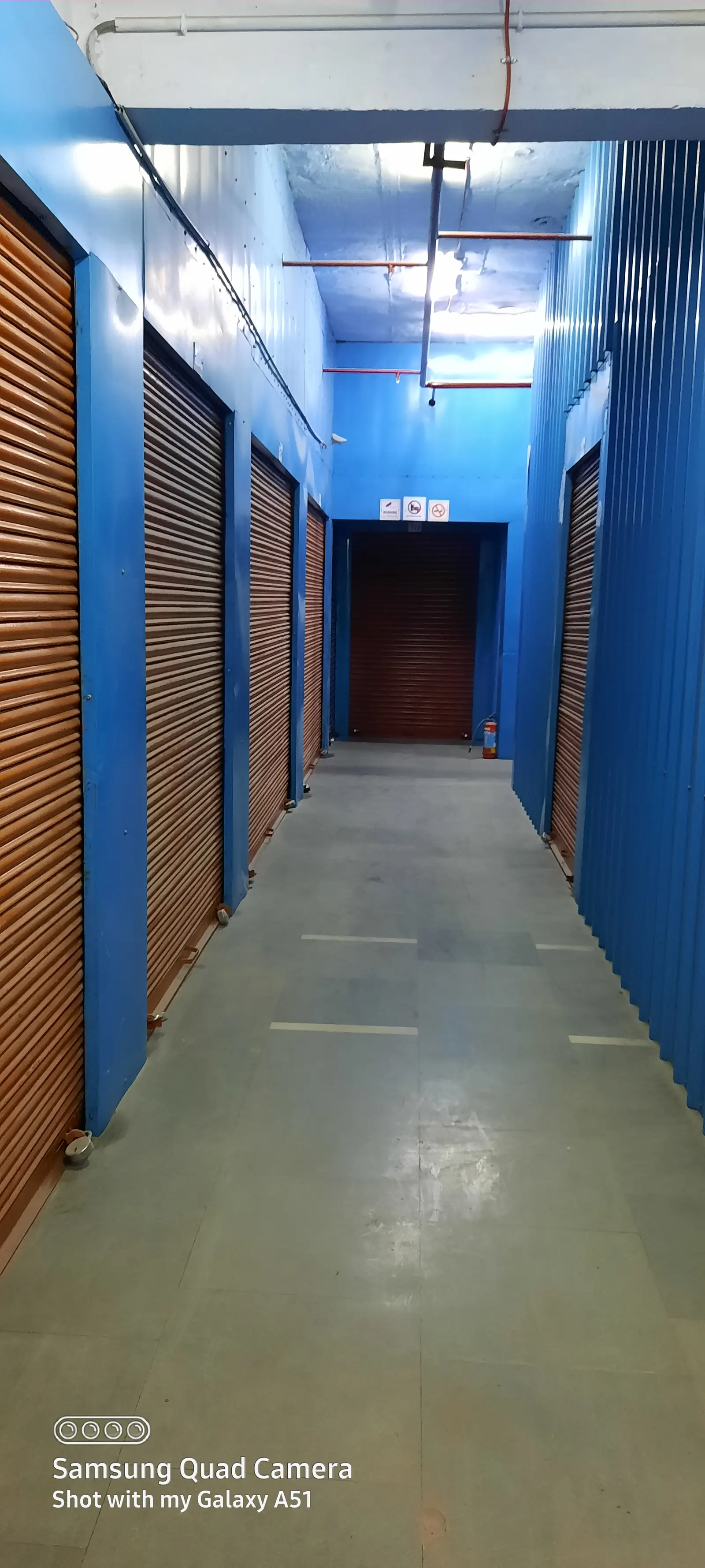 A long storage corridor with blue walls and lockable rolling-shutter units on both sides, and a fire extinguisher at the far end