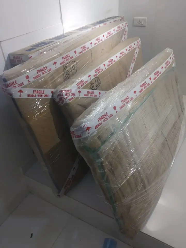 Three large flat cartons wrapped in stretch film with FRAGILE HANDLE WITH CARE tape along every edge, leaning against a wall
