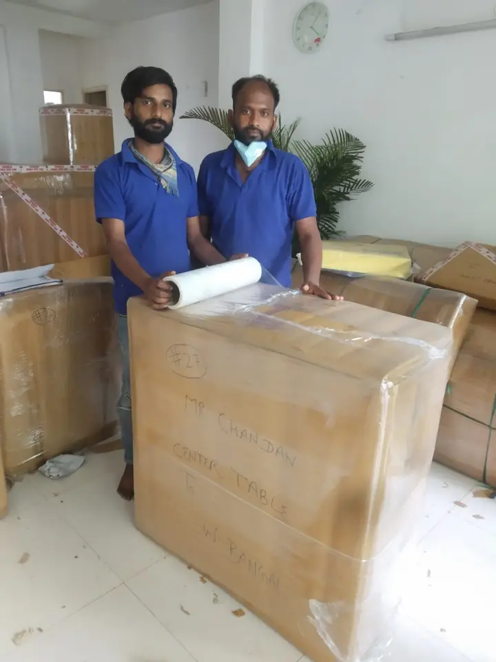 Two crew members in blue shirts wrapping stretch film around a carton labelled “centre table, to West Bengal”, numbered 27