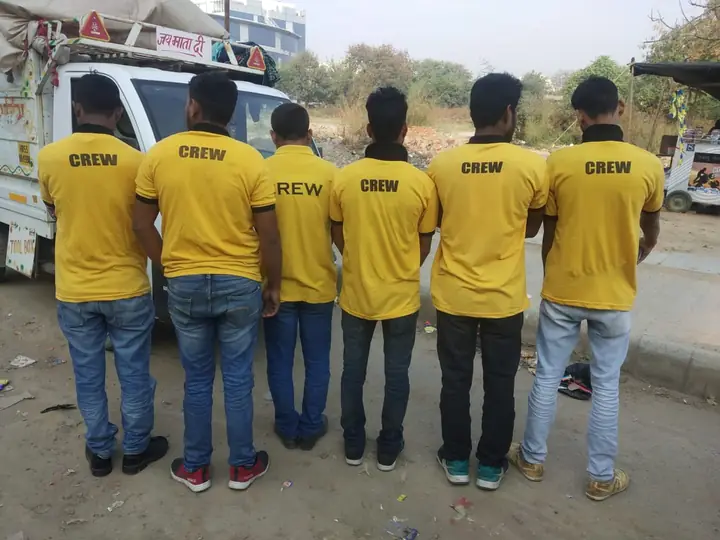 Six crew members in yellow CREW T-shirts, seen from behind, standing beside a small loading vehicle