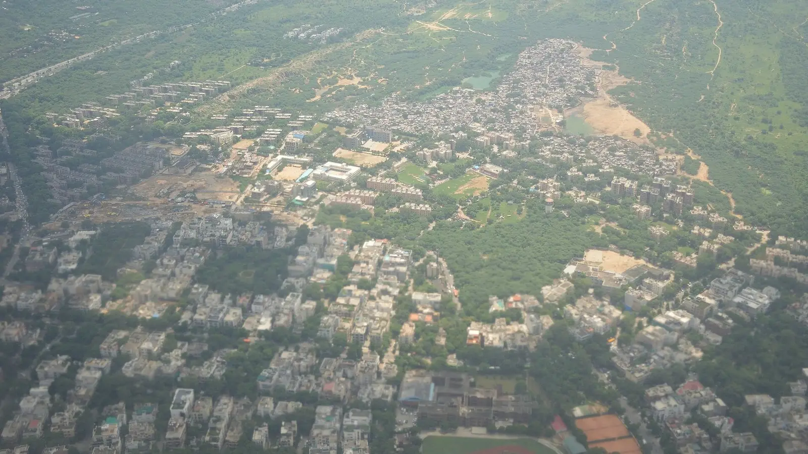 Illustration: aerial view of Vasant Vihar's low-rise bungalows and sports grounds with the ridge forest beyond (hazy).